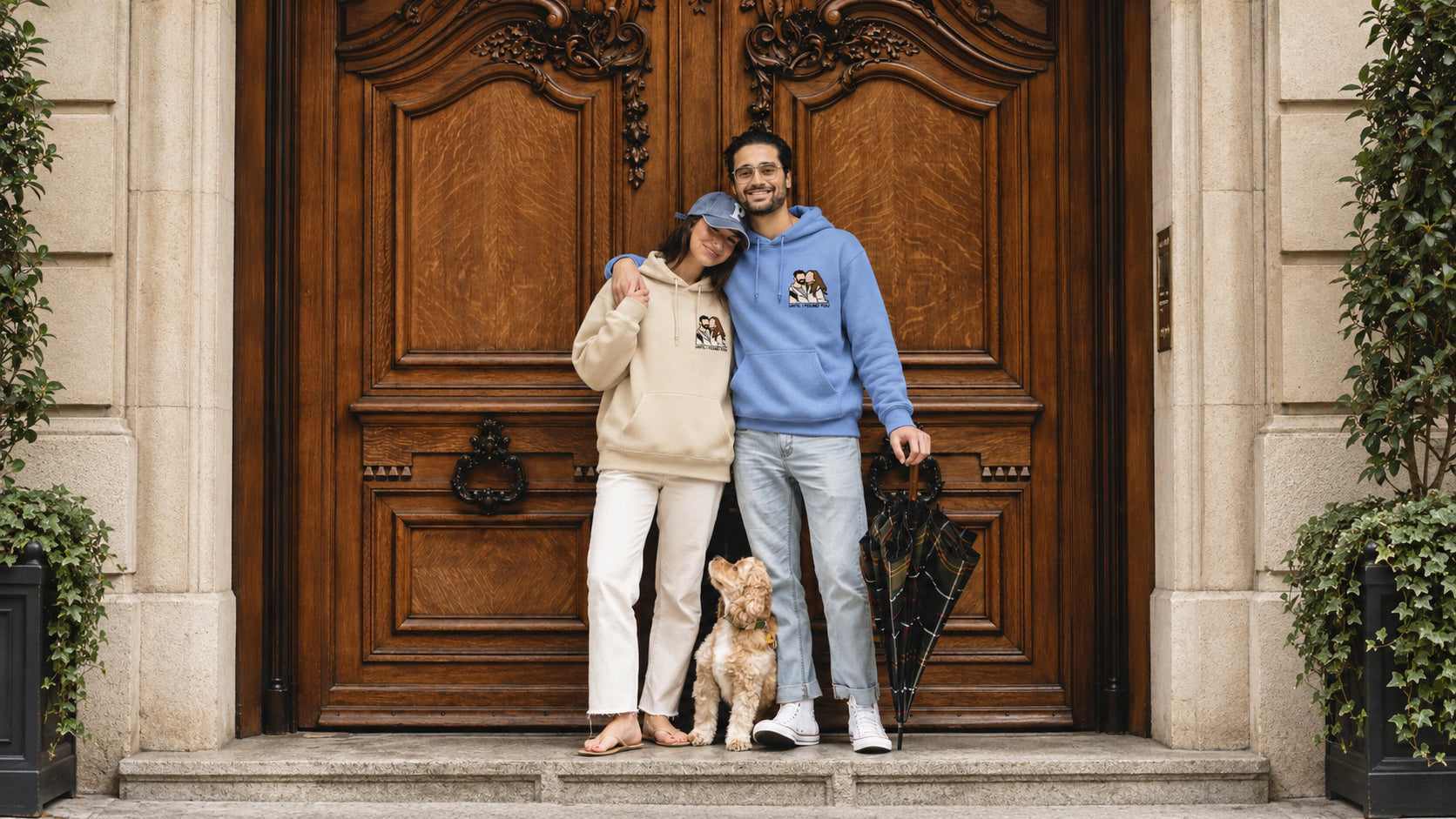 A couple wearing custom embroidered hoodies with their cat outside a wooden doorway